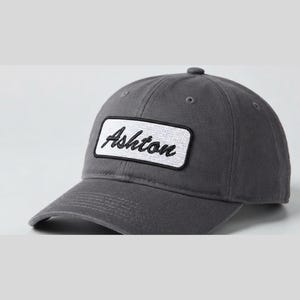 May include: A charcoal grey baseball cap featuring a rectangular white patch on the front panel. The patch contains the word Ashton embroidered in black cursive script. The cap has a curved brim, stitched eyelets, and a button on the crown.