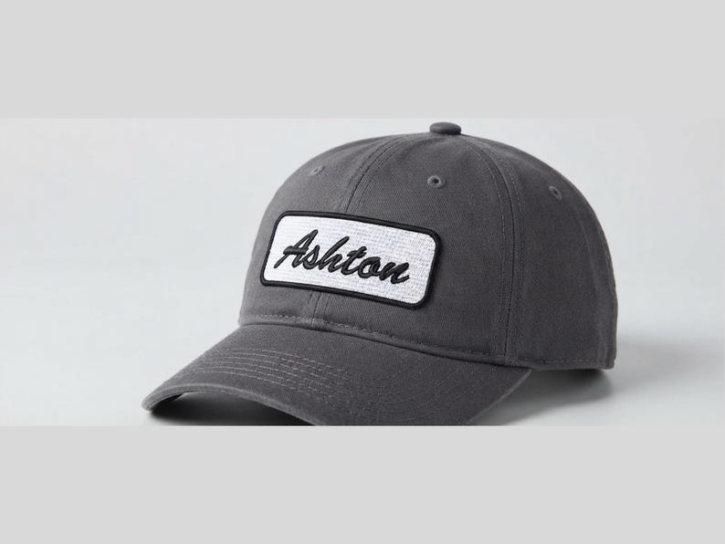 May include: A charcoal grey baseball cap featuring a rectangular white patch on the front panel. The patch contains the word Ashton embroidered in black cursive script. The cap has a curved brim, stitched eyelets, and a button on the crown.
