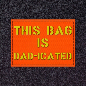 May include: An orange rectangular patch with the text "THIS BAG IS DAD-ICATED" in yellow, stitched onto a dark grey, textured background. The text is in a bold, sans-serif font.