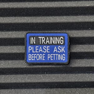 Könnte beinhalten: Ein rechteckiger blauer Aufnäher mit weiß gesticktem Text "IN TRAINING PLEASE ASK BEFORE PETTING". Der Aufnäher hat einen schwarzen Rand und befindet sich auf einem strukturierten schwarzen Hintergrund.