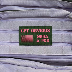 May include: A close-up of a purple tactical backpack with a green patch. The patch features the text "CPT OBVIOUS" above "NKDA A POS" in pink, along with a pink American flag.