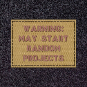 May include: A tan rectangular patch with the text "WARNING: MAY START RANDOM PROJECTS" in pinkish-brown. The patch has a black dashed border, set against a dark grey, textured background.