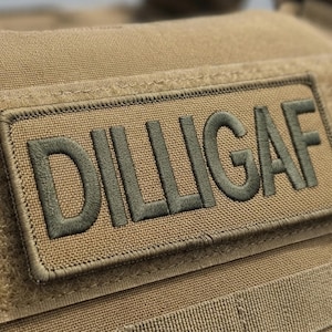 May include: A rectangular tan tactical patch featuring the embroidered text DILLIGAF in dark olive green thread. The patch is securely attached to a tan fabric surface with a hook-and-loop fastener backing, displaying a clean stitched border.