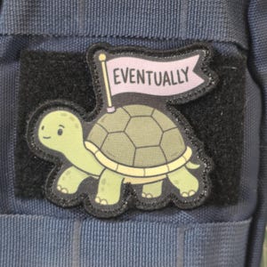 May include: A rectangular morale patch featuring a light green turtle with a dark green shell. The turtle carries a light purple flag on its back with the word EVENTUALLY printed in black capital letters. The patch has a black border.