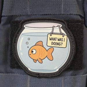 May include: A circular fabric patch with a black border featuring a cartoon goldfish swimming in a round glass bowl. A small rectangular sign hangs from the rim of the bowl with the text WHAT WAS I DOING? printed in black capital letters.