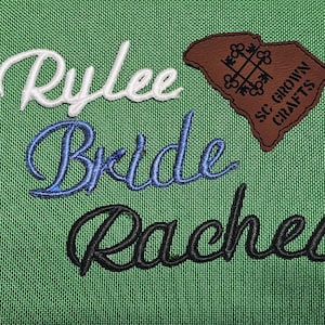 Könnte beinhalten: Grüner Stoff mit gestickten Namen: "Rylee" in Weiß, "Bride" in Blau und "Rachel" in Schwarz. Ein braunes Lederpatch mit dem Text "SC GROWN CRAFTS" und einer Umrisszeichnung des Bundesstaates ist ebenfalls vorhanden.