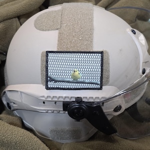 May include: A tan tactical helmet featuring a rectangular patch with a black border. The patch displays a small yellow bird perched on a branch against a background filled with the repeated letter A in a grid pattern, creating a textured effect.