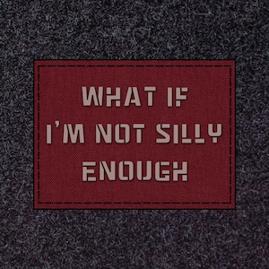 May include: A rectangular patch with the text "WHAT IF I'M NOT SILLY ENOUGH" in a light beige colour. The patch is a deep red with a black border and is set against a dark grey, textured background.