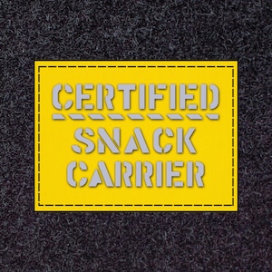 May include: A yellow rectangle with the text "CERTIFIED SNACK CARRIER" in grey, stencil-style lettering. The rectangle has a black dashed border, set against a dark grey, textured background.