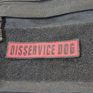 Pode incluir: Um emblema retangular com fundo vermelho e borda preta fixado em uma superfície de tecido preto. O emblema apresenta o texto "DISSERVICE DOG" em letras maiúsculas pretas, em negrito e sem serifa. É fixado com um fecho de velcro.