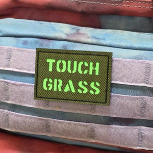 May include: A rectangular patch with the words "TOUCH GRASS" in bright green stencil-style lettering. The patch has a dark green background and a black border. The patch is placed on a colourful, textured background.