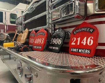 May include: Four red, black, and wooden firefighter helmet plaques with white lettering. The plaques read "Engineer 535 4093 Fort Myers", "Firefighter 5906 Fort Myers", "Engineer 2146 Fort Myers", and "Engineer 535 4093 Fort Myers".