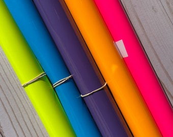 Op de afbeelding: Zes rollen vinyl-kleefolie in verschillende kleuren, waaronder neon geel, blauw, paars, oranje en roze. De rollen zijn op elkaar gestapeld en worden bij elkaar gehouden met een elastiekje.