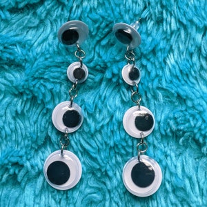 May include: A pair of dangle earrings with three googly eyes on each earring. The eyes are white with black pupils and are connected by silver chains.