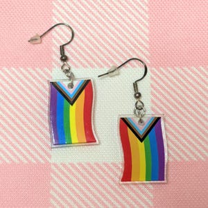 Op de afbeelding: Een paar hangende oorbellen met rechthoekige hangers in het ontwerp van de Progress Pride-vlag. De vlag bevat verticale strepen in rood, oranje, geel, groen, blauw en paars, met een chevronpatroon in zwart, bruin, lichtblauw, roze en wit.