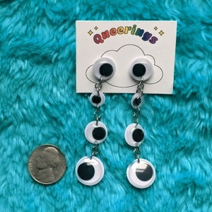 May include: A pair of dangling earrings with a silver chain and four white googly eyes with black pupils. The earrings are on a white card with the text "queering" in black.