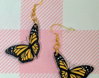 May include: A pair of gold-colored drop earrings featuring monarch butterfly designs. The earrings have yellow and black wings with white dot details. The background is a pink and white checkered pattern.
