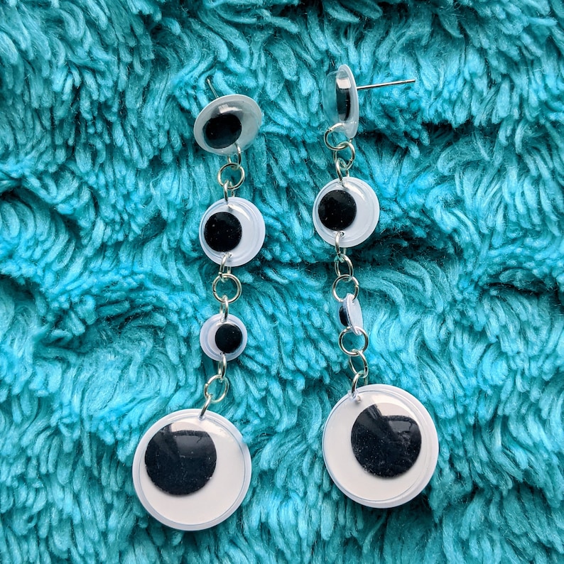 May include: A pair of white and black eye-shaped earrings with silver chains. Each earring has three white circles with black centers.
