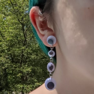 May include: A pair of dangle earrings with a silver chain and multiple white plastic eyeballs. The earrings are hanging from the earlobe.