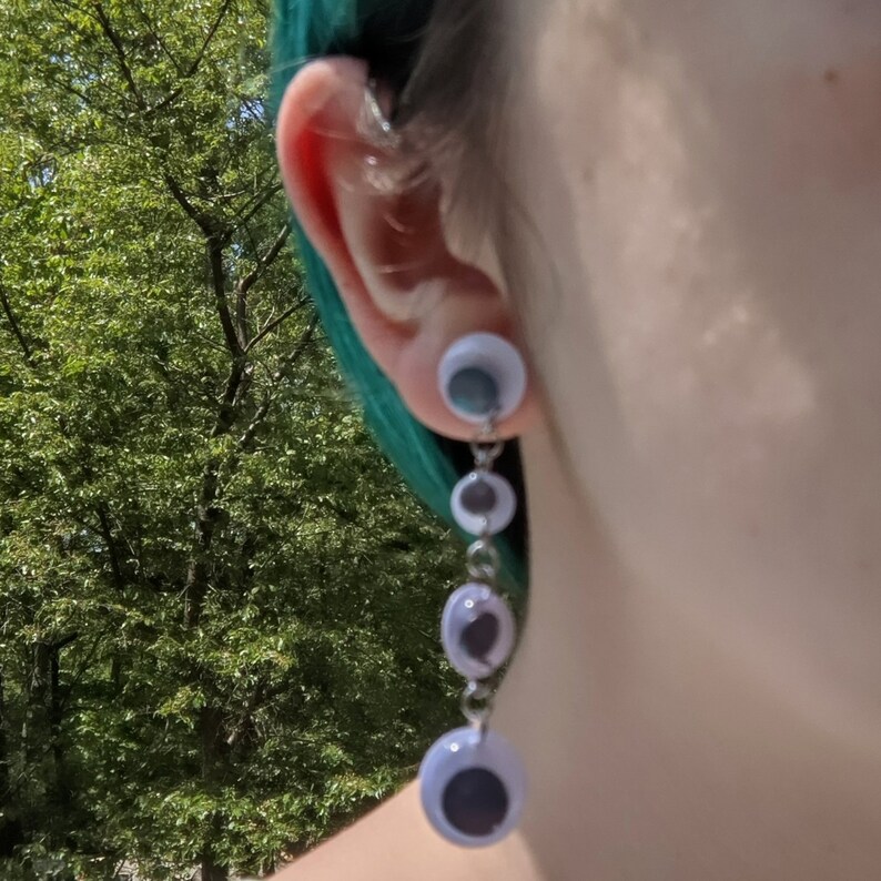 May include: A pair of dangle earrings with a silver chain and multiple white plastic eyeballs. The earrings are hanging from the earlobe.