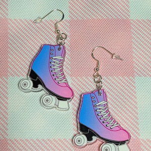 May include: A pair of roller skate earrings with a pink and blue ombre design. The earrings are made of acrylic and have silver hooks.