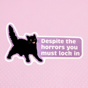 May include: A sticker featuring a black cat with its tongue sticking out, outlined in white and purple. The sticker has the text "Despite the horrors you must lock in" in white on a purple background, set against a pink polka-dot backdrop.