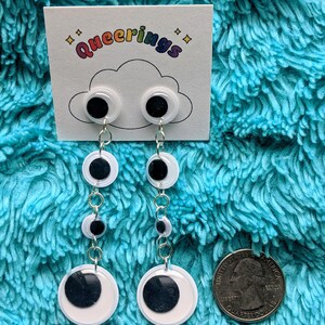 May include: A pair of white and black googly eye dangle earrings with silver chains. The earrings are displayed on a white card with the text "queering" in rainbow letters.