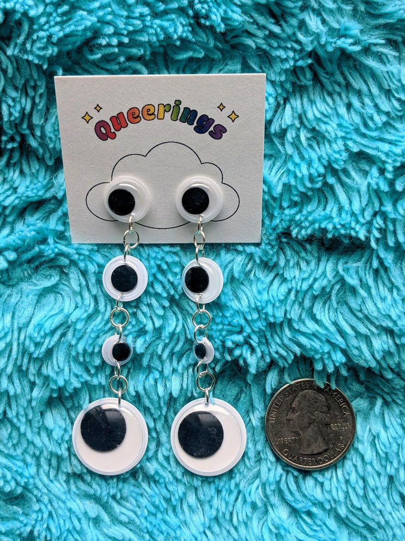 May include: A pair of white and black googly eye dangle earrings with silver chains. The earrings are displayed on a white card with the text "queering" in rainbow letters.