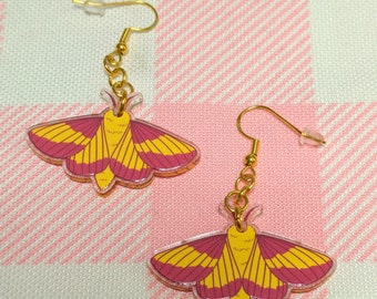 Puede incluir: Un par de pendientes de oro con un diseño de polilla rosa y amarilla. Las polillas tienen una apariencia translúcida y están acentuadas con detalles dorados.