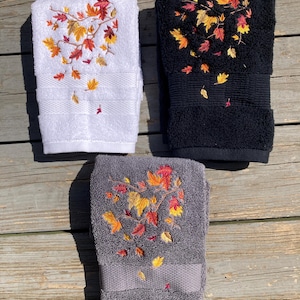 May include: Three embroidered hand towels in white, black, and gray. Each towel features a detailed embroidery of autumn leaves in shades of red, orange, and yellow. The towels are folded and displayed on a wooden surface.