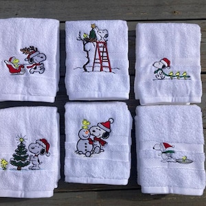 Puede incluir: Seis toallas de mano blancas con diseños navideños bordados. Cada toalla presenta a Snoopy en varias escenas navideñas, incluyendo un trineo, una escalera y un muñeco de nieve. Las toallas están dobladas y apiladas, mostrando el bordado festivo.