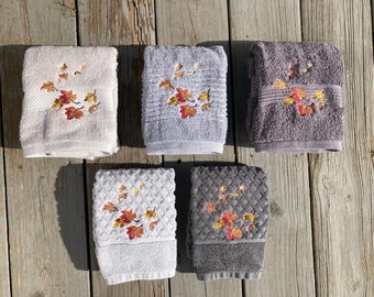 Puede incluir: Conjunto de seis toallas con hojas de otoño bordadas en tonos naranja, amarillo y marrón. Las toallas son de dos estilos diferentes, una con tejido de gofre y la otra con textura lisa. Las toallas son de dos colores, gris y blanco.