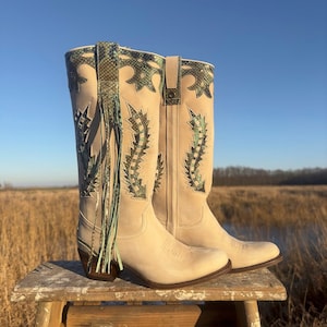 May include: A pair of off-white cowboy boots with green and white patterned embroidery and a green fringe detail.