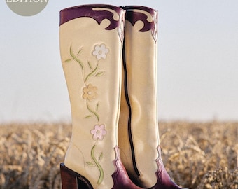 Puede incluir: Un par de botas de vaquero de tacón alto, color crema, con detalles burdeos y bordados florales. Las botas se exhiben sobre un taburete de madera, con un fondo de trigo. Las palabras "EDICIÓN LIMITADA" están en un círculo en la esquina superior izquierda.