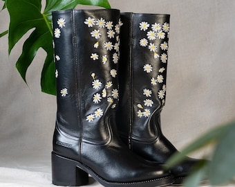 Könnte beinhalten: Schwarze Leder-Cowgirl-Stiefel mit hohem Schaft und Blockabsatz. Die Stiefel sind mit weißen Gänseblümchen-Stickereien verziert.