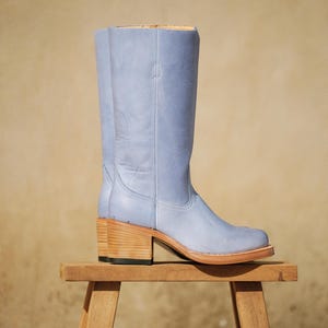 Könnte beinhalten: Ein hellblauer Leder-Cowboystiefel mit einem Holzabsatz. Der hohe Stiefel wird auf einem kleinen Holzschemel vor einem neutralen Hintergrund präsentiert. Der Stiefel hat eine runde Spitze und eine glatte Oberfläche.