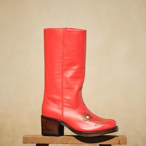 Może przedstawiać: Żywy czerwony skórzany but z kwiatowym wzorem. But ma kwadratowy nosek, obcas w kształcie bloku i brązową aplikację kwiatową. But ma około 30 cm wysokości i jest prezentowany na małym drewnianym stołku.