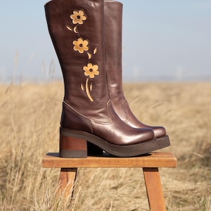 Może przedstawiać: Brązowe skórzane kozaki do kolan z platformą i obcasem w kształcie klocka. Buty mają kwiatowy wzór w jaśniejszym brązie. Buty stoją na małym drewnianym stołku na polu.