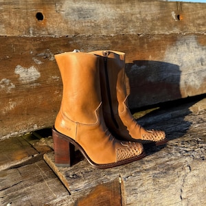May include: A pair of tan leather cowboy boots with a square toe and a stacked wooden heel. The boots have a snakeskin pattern on the toe and a zipper closure. The boots are approximately 25 cm tall.