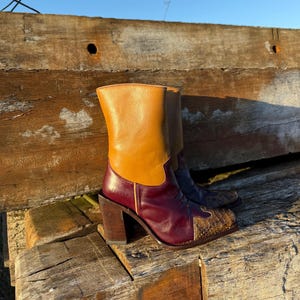 Może przedstawiać: Skórzane botki z grubym obcasem. Buty mają wzór color block z beżem, bordowym i wzorem przypominającym skórę węża na nosku. Ustawione na rustykalnym drewnianym tle pod błękitnym niebem.