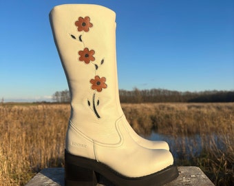 May include: White leather knee-high boots with a floral design featuring brown flowers. The boots have a chunky black heel and a platform sole. The boots are sitting on a wooden stool. The brand name "SENDRA" is visible on the side of the boot.