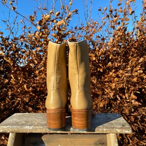 May include: A pair of tan leather boots with a block heel. The boots are sitting on a wooden stool against a backdrop of brown leaves and a blue sky.