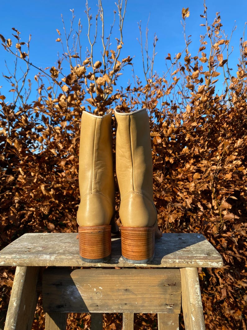 May include: A pair of tan leather boots with a block heel. The boots are sitting on a wooden stool against a backdrop of brown leaves and a blue sky.