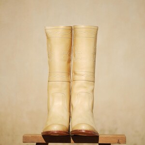 May include: A pair of tall, light tan leather boots with decorative stitching. The boots are knee-high, with a rounded toe and a brown sole. They are displayed on a small wooden stool against a neutral background.