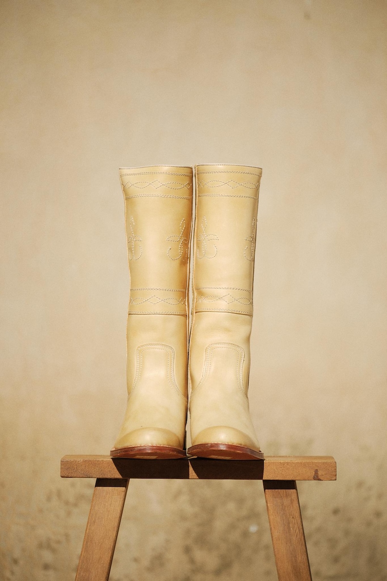 May include: A pair of tall, light tan leather boots with decorative stitching. The boots are knee-high, with a rounded toe and a brown sole. They are displayed on a small wooden stool against a neutral background.