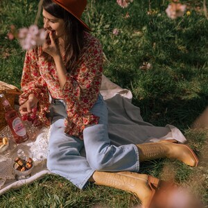 Puede incluir: Una escena de picnic con una persona que lleva un sombrero color &oacute;xido, una blusa floral y unos vaqueros azul claro. La escena incluye una botella de limonada rosa, aperitivos y botas de vaquero altas de color marr&oacute;n claro.