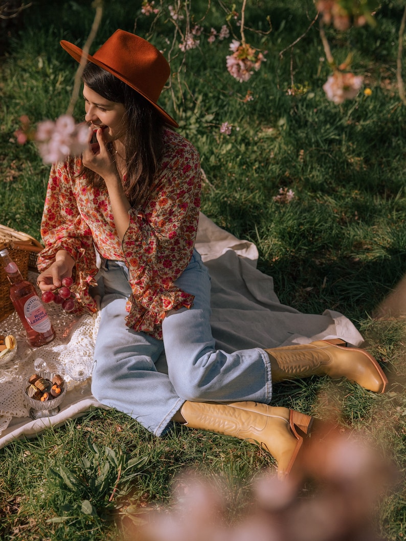 Puede incluir: Una escena de picnic con una persona que lleva un sombrero color &oacute;xido, una blusa floral y unos vaqueros azul claro. La escena incluye una botella de limonada rosa, aperitivos y botas de vaquero altas de color marr&oacute;n claro.