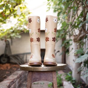 May include: A pair of beige leather boots with floral embroidery. The boots are sitting on a wooden stool.