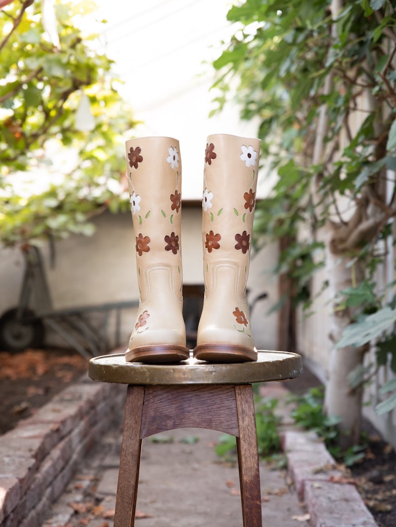 May include: A pair of beige leather boots with floral embroidery. The boots are sitting on a wooden stool.