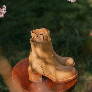 May include: Tan leather ankle boots with a chunky heel and a side zip. The boots are sitting on a wooden stool. The brand name "SENDRA" is visible on the side of the boot.